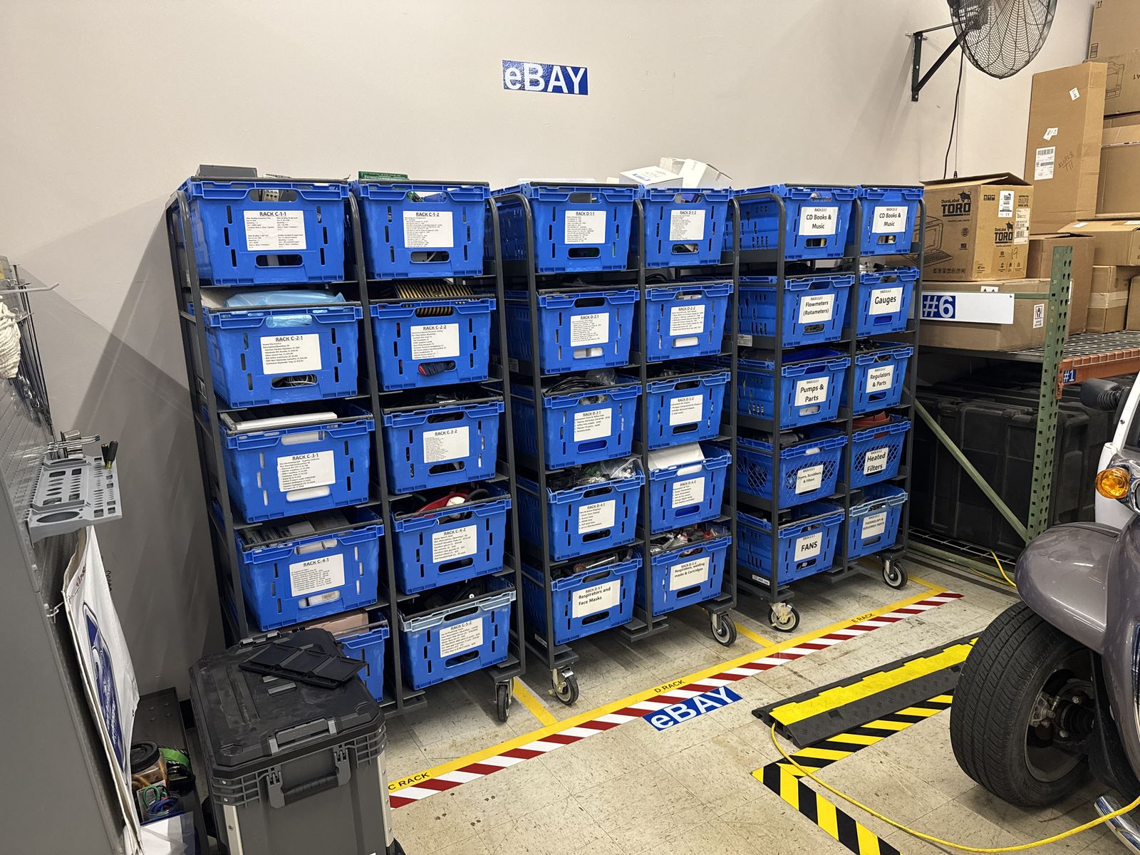 The eBay packing bay: rows of stacked blue parts bins under a sign reading eBay, with a hazard-striped floor marking