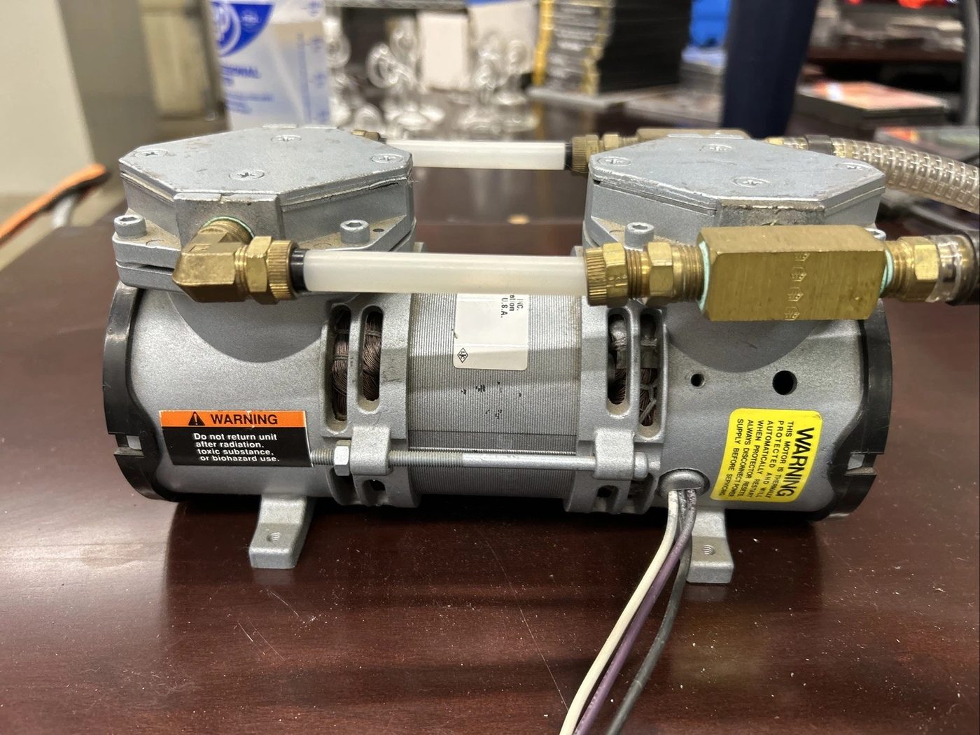 A twin-head diaphragm pump with brass fittings and a warning label, sitting on a workbench
