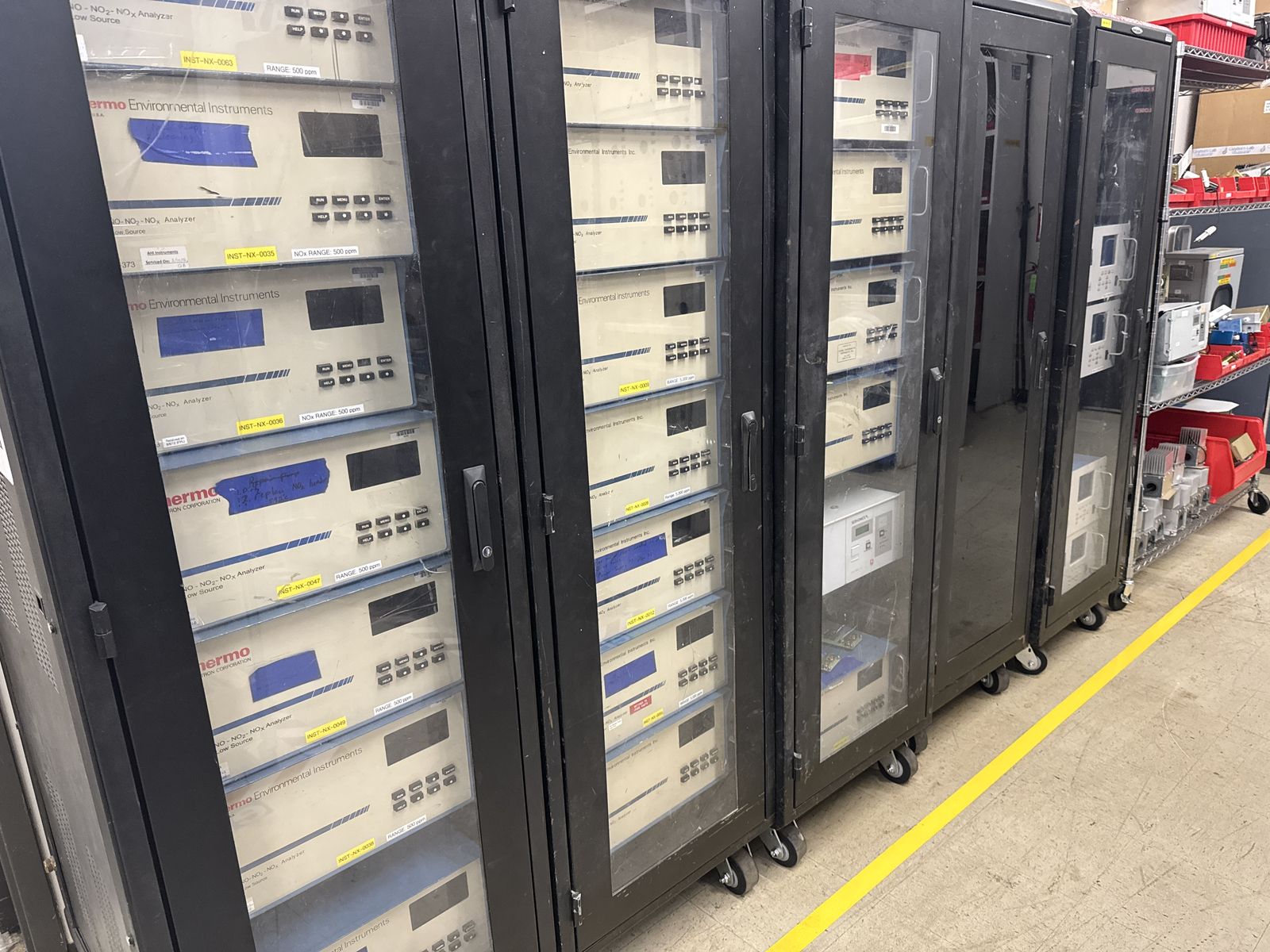 Rows of nineteen-inch equipment racks filled with Thermo gas analyzers on a warehouse floor, aisle markings painted in yellow